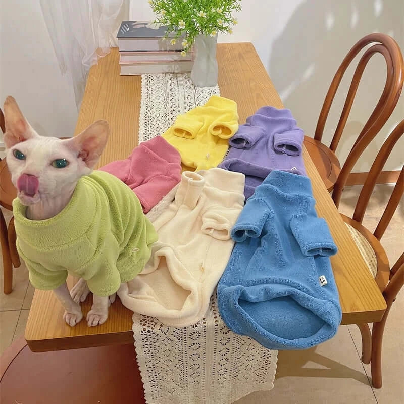 Sphynx cat wearing a green fleece coat with various colorful fleece coats on a wooden table.