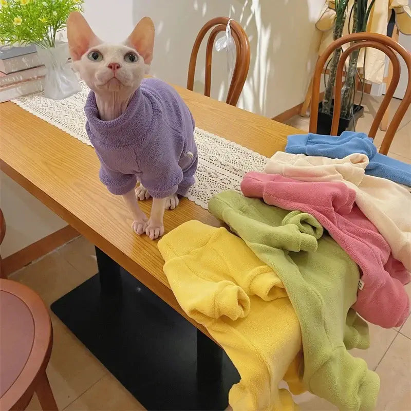 Sphynx cat wearing a purple fleece coat, surrounded by colorful fleece sweatshirts on a wooden table.