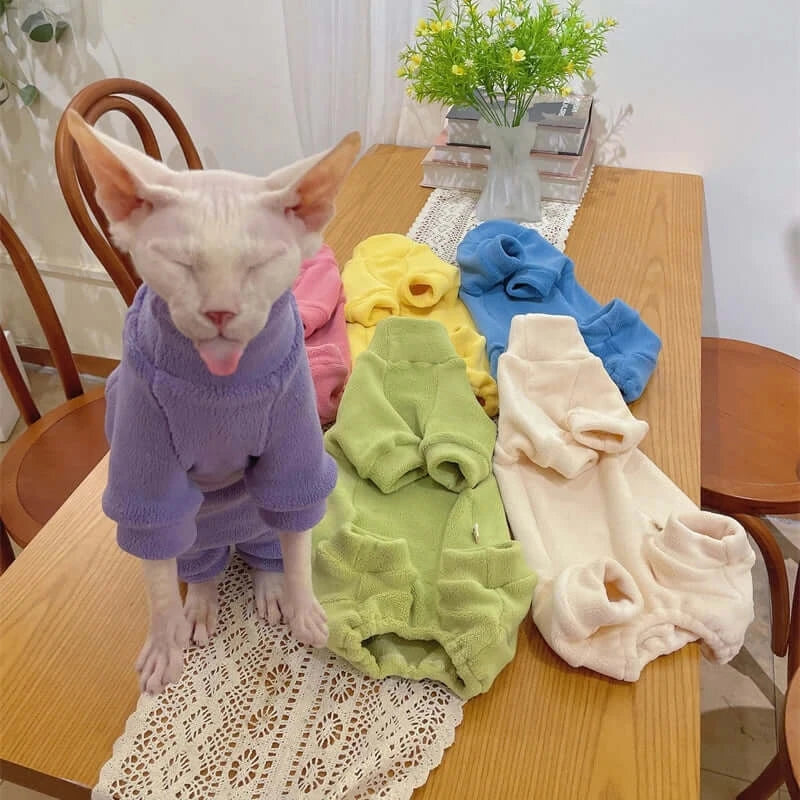 Sphynx cat in warm fleece coat among colorful sweatshirts on a wooden table, showcasing autumn/winter pet fashion.
